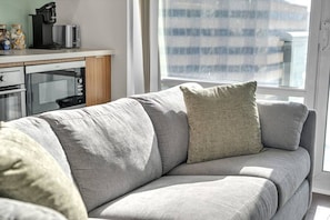 Ein modernes Wohnzimmer mit einem grauen Sofa, einer Kaffeemaschine und einem Fenster mit Jalousien.