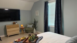 A bedroom with a bed, a TV, a small side table, a chair, and a window with curtains. - Peak-A-Blue: Beautifully Renovated Oceanfront Home with Hot Tub (Oak Island)