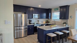 A modern kitchen with dark cabinetry, stainless steel appliances, a central island with bar stools, and a window with blinds. - Peak-A-Blue: Beautifully Renovated Oceanfront Home with Hot Tub (Oak Island)