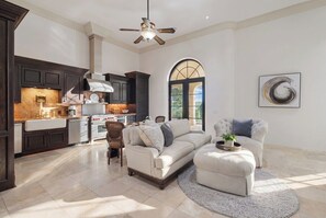 A spacious living area with a sofa and ottoman, a dining table, and a kitchen with dark cabinetry and a large window. - Blanco River estate + 2 pools + court (Wimberly)