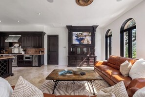 A spacious living area with a leather sofa, a coffee table with books and remote controls, and a kitchen with dark cabinetry and a large window. - Blanco River estate + 2 pools + court (Wimberly)