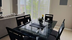 A modern living room with a glass dining table, black chairs, a grey sofa, and a large window with vertical blinds. - Hope Oasis (Kingston)
