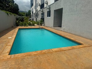 Une piscine rectangulaire bordée d’une terrasse en bois, entourée d’un petit muret et de verdure.