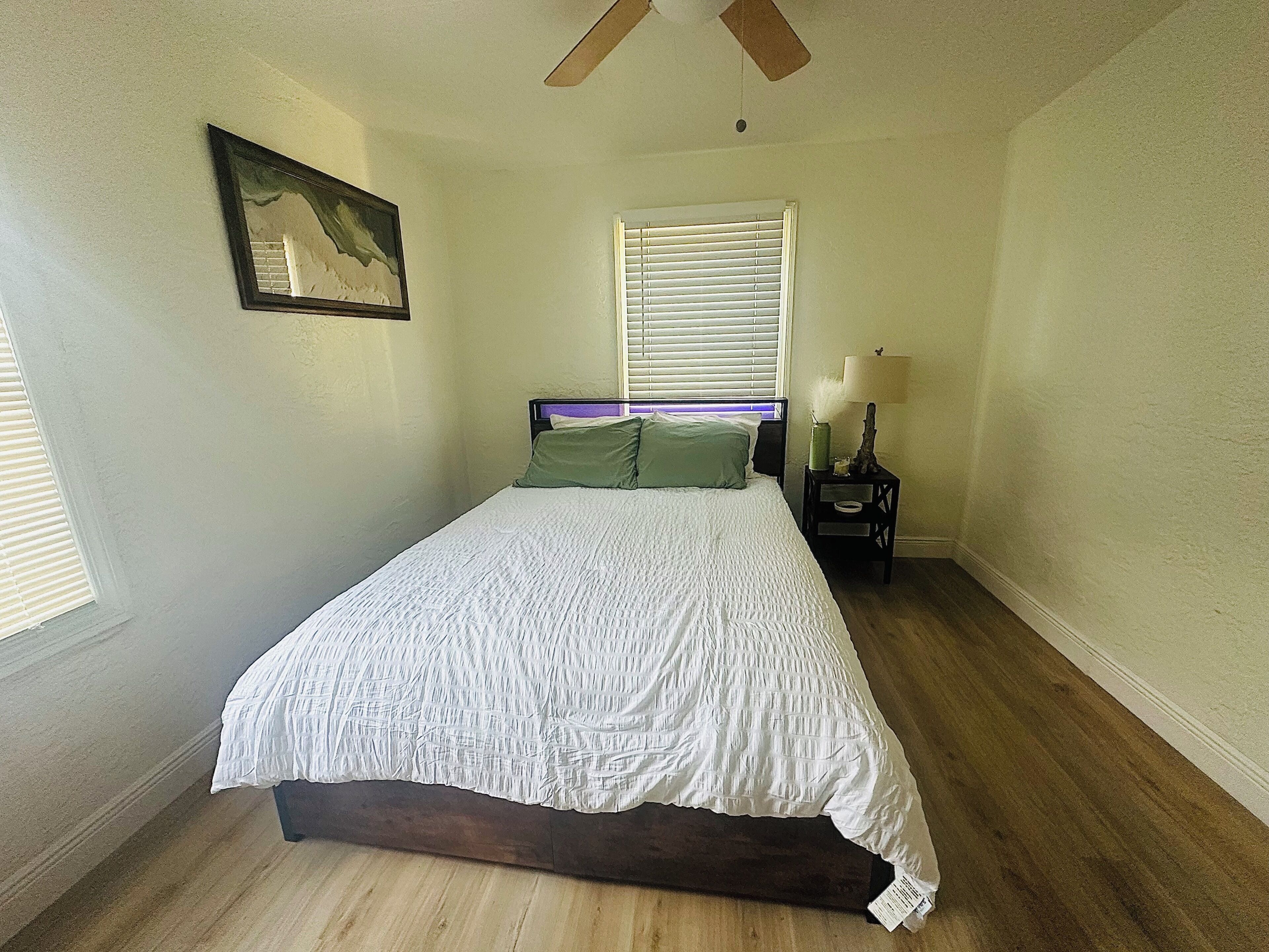 Une chambre à coucher comprenant un lit, une table de chevet, un ventilateur de plafond, une fenêtre avec des stores et un tableau accroché au mur.