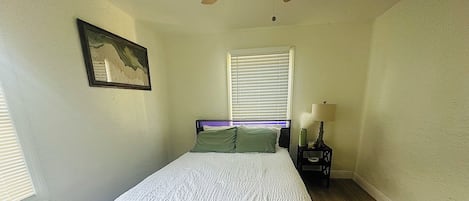 Un dormitorio con cama, mesita de noche, ventilador de techo, ventana con persianas y un cuadro colgado en la pared.