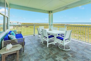 Een overdekt terras met een tafel, stoelen en een bank met kussens, en uitzicht op het strand.