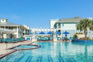 A swimming pool with lounge chairs and umbrellas, surrounded by buildings with balconies and a palm tree. - Beach Baby Bungalow - Beachfront Condo with Gulf Views & Pointe West Amenities (Galveston)