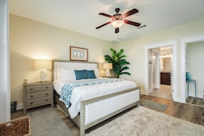 A bedroom with a bed, nightstands, a ceiling fan, a potted plant, and a view into the bathroom. - Beach Baby Bungalow - Beachfront Condo with Gulf Views & Pointe West Amenities (Galveston)