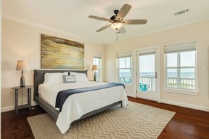 Una camera da letto con un letto, una ventola a soffitto, un quadro e vista sull'oceano attraverso ampie finestre.