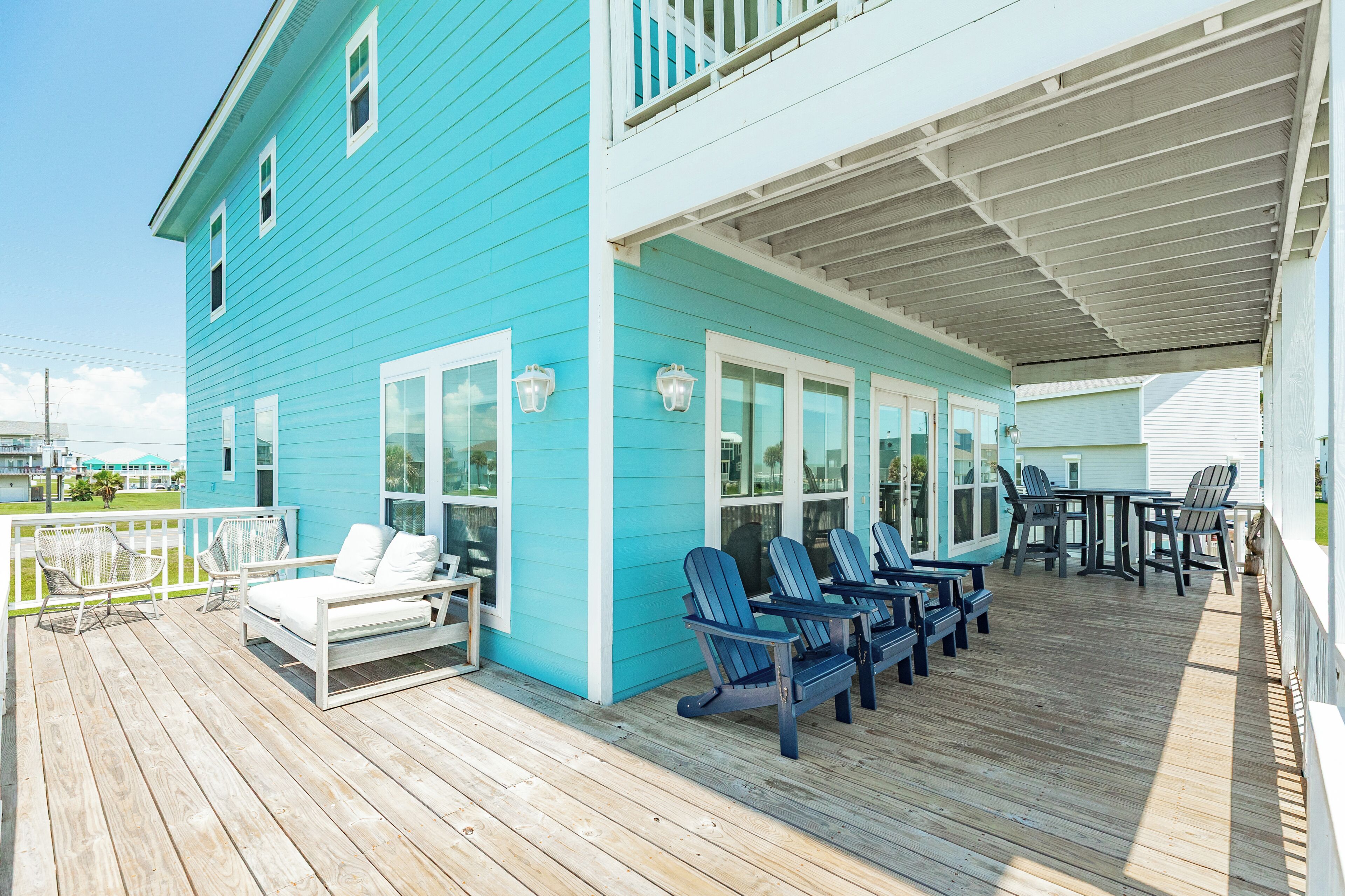A covered deck with Adirondack chairs and a white sofa, a wooden floor, and a turquoise building with white trim.