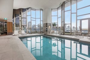 An indoor swimming pool with clear views of the cityscape through large windows, surrounded by lounge chairs and a small table. - Aerie at West Side City View and Wellness Facilities (Melbourne)