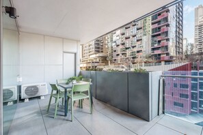 Varanda com mesa e cadeiras, aparelhos de ar condicionado e vista de um edifício de vários andares.