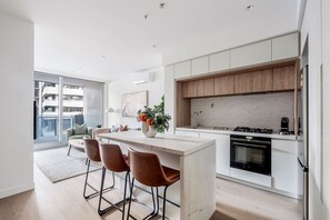 A modern kitchen with a central island, bar stools, a built-in oven, and a view of a residential building. - Aerie at West Side City View and Wellness Facilities (Melbourne)
