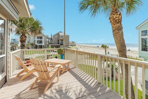Eine Holzterrasse mit Gartenmöbeln, mit Blick auf einen Strand und Wohnhäuser.