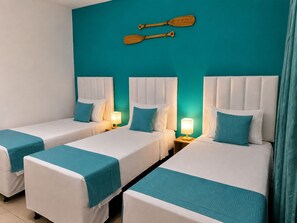 Una habitación de hotel con tres camas, cada una con cabecero blanco y ropa de cama color turquesa. Hay dos palas de madera fijadas a la pared sobre las camas.