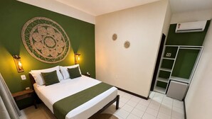 Quarto de hotel com cama, parede verde, um objeto decorativo na parede, tapete pequeno e área de cozinha compacta.
