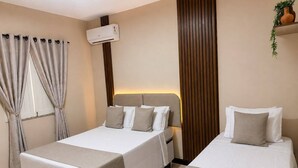 Quarto de hotel com duas camas, janela com cortinas, ar condicionado e uma planta numa prateleira.