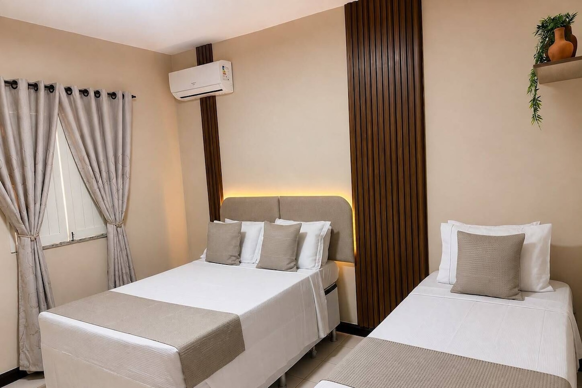 A hotel room with two beds, a window with curtains, an air conditioning unit, and a plant on a shelf.