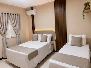 A hotel room with two beds, a window with curtains, an air conditioning unit, and a plant on a shelf. - Pousada Lagoas de Guarajuba (Camaçari)