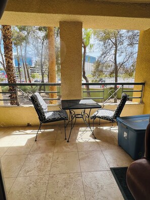Ein Balkon mit Tisch und zwei gemusterten Stühlen, einer blauen Aufbewahrungsbox und Blick auf eine Straße mit Bäumen und Gebäuden.