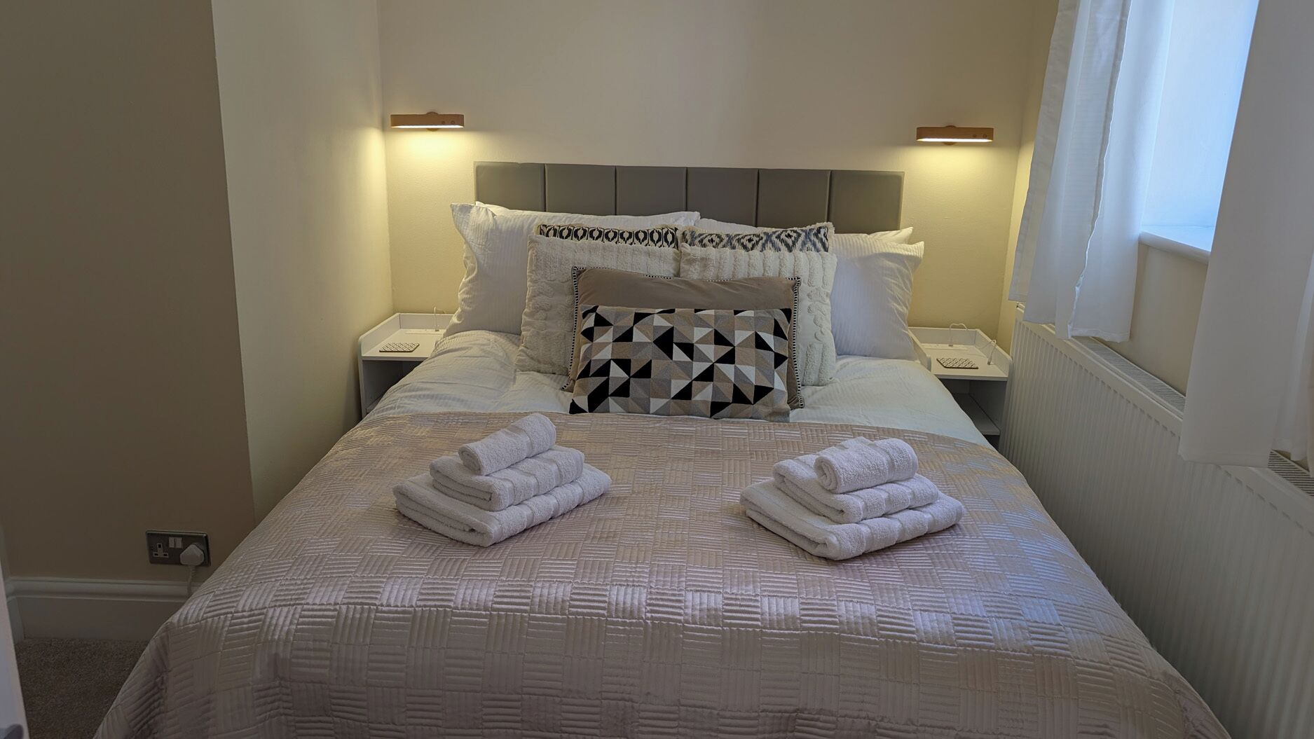 A neatly made bed with a patterned bedspread and pillows, two folded towels on the bed, bedside lamps, and a window with curtains.