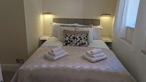 Ein ordentlich bezogenes Bett mit gemustertem Bettbezug und Kissen, zwei gefaltete Handtücher auf dem Bett, Nachttischlampen und ein Fenster mit Vorhängen.