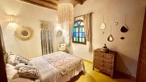 A bedroom with a bed, a dresser, a hanging light fixture, a window with curtains, and wall decor. - Villa under the trade winds (Essaouira)