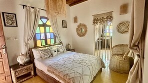 Ein Schlafzimmer mit einem Bett, einem Nachttisch, einem Ventilator, einem Korbstuhl und einem Fenster mit Vorhängen.