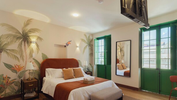 A hotel room with a bed, a nightstand, a lamp, a mirror, and a view of the outside through green doors.