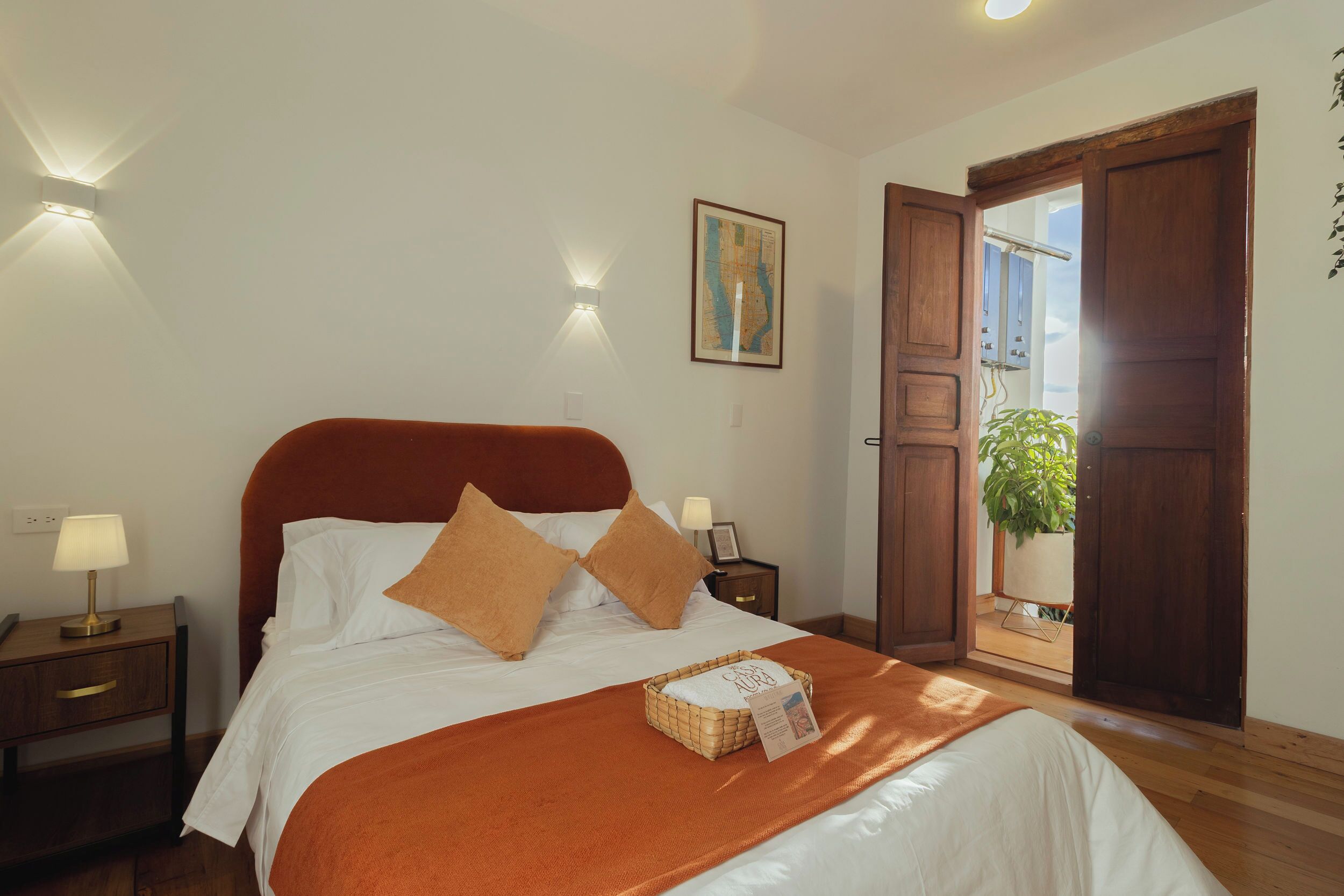Un lit bien fait, avec une tête de lit marron, du linge de lit blanc et deux oreillers marron. On y trouve une table de chevet avec une lampe, une carte encadrée accrochée au mur et une porte en bois donnant sur l’extérieur.