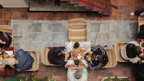A group of people seated on woven mats around a central table with food, in a room with tiled flooring and a brick wall.