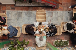 Un grupo de personas sentadas sobre esteras tejidas alrededor de una mesa central con comida, en una habitación con suelo de baldosas y pared de ladrillo.