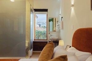 A hotel room with a bed, a bathroom with a sink and mirror, and a view of a building through a window.