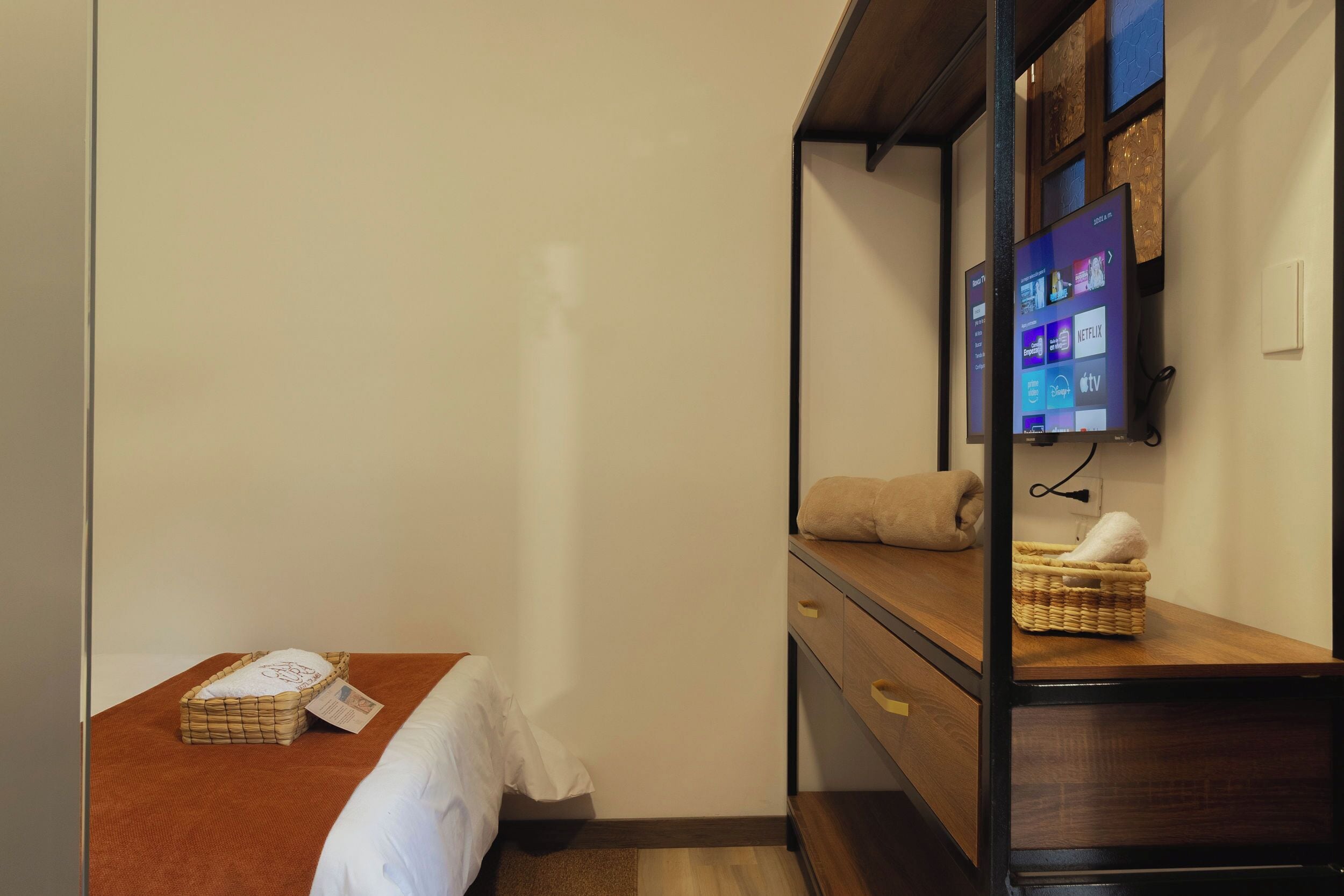 Una habitación de hotel con una cama, una mesita de noche de madera, un televisor montado en la pared y una pequeña cesta de mimbre sobre la mesita.