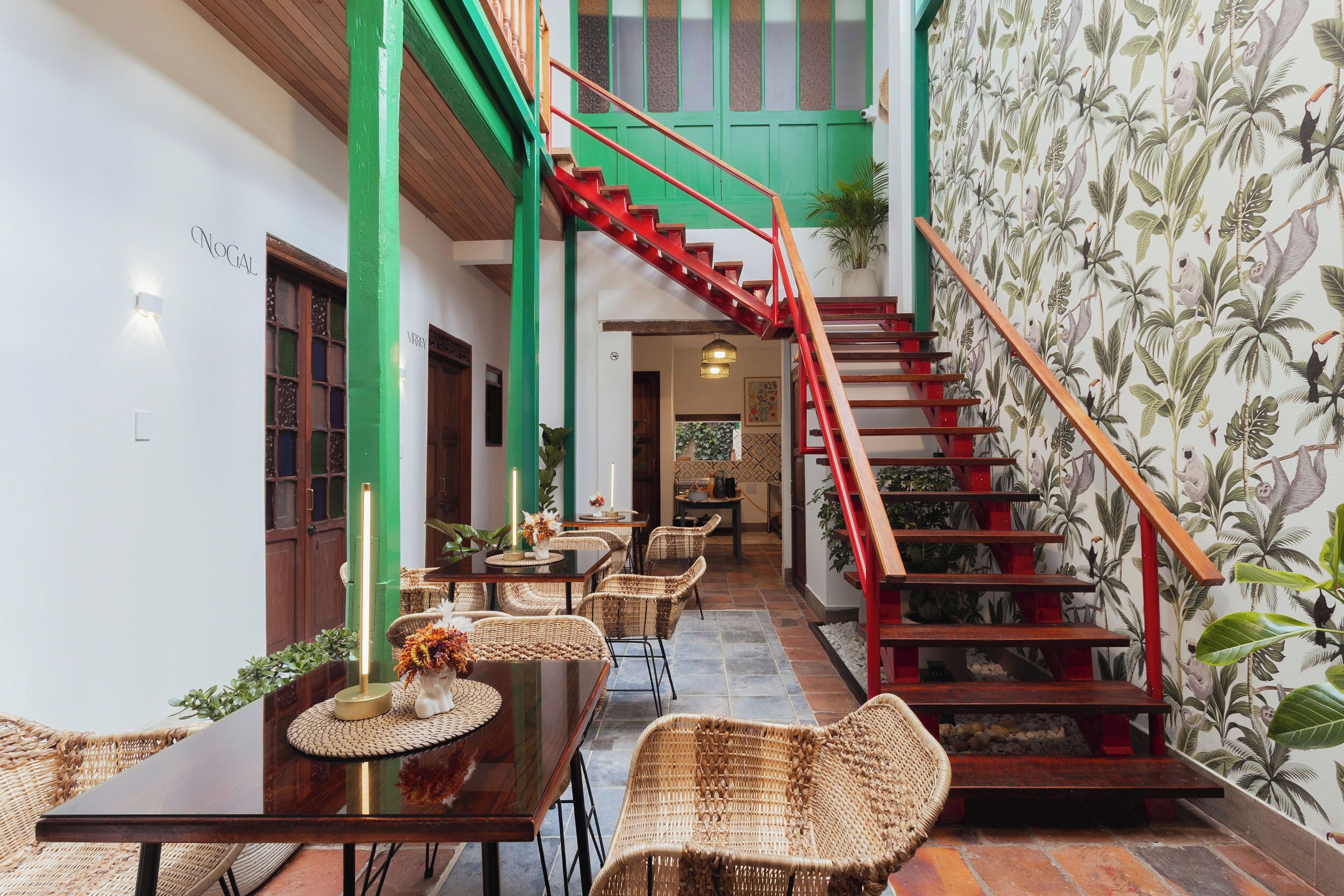 A dining area with wicker furniture, a staircase with red railings, and a tropical wallpaper.