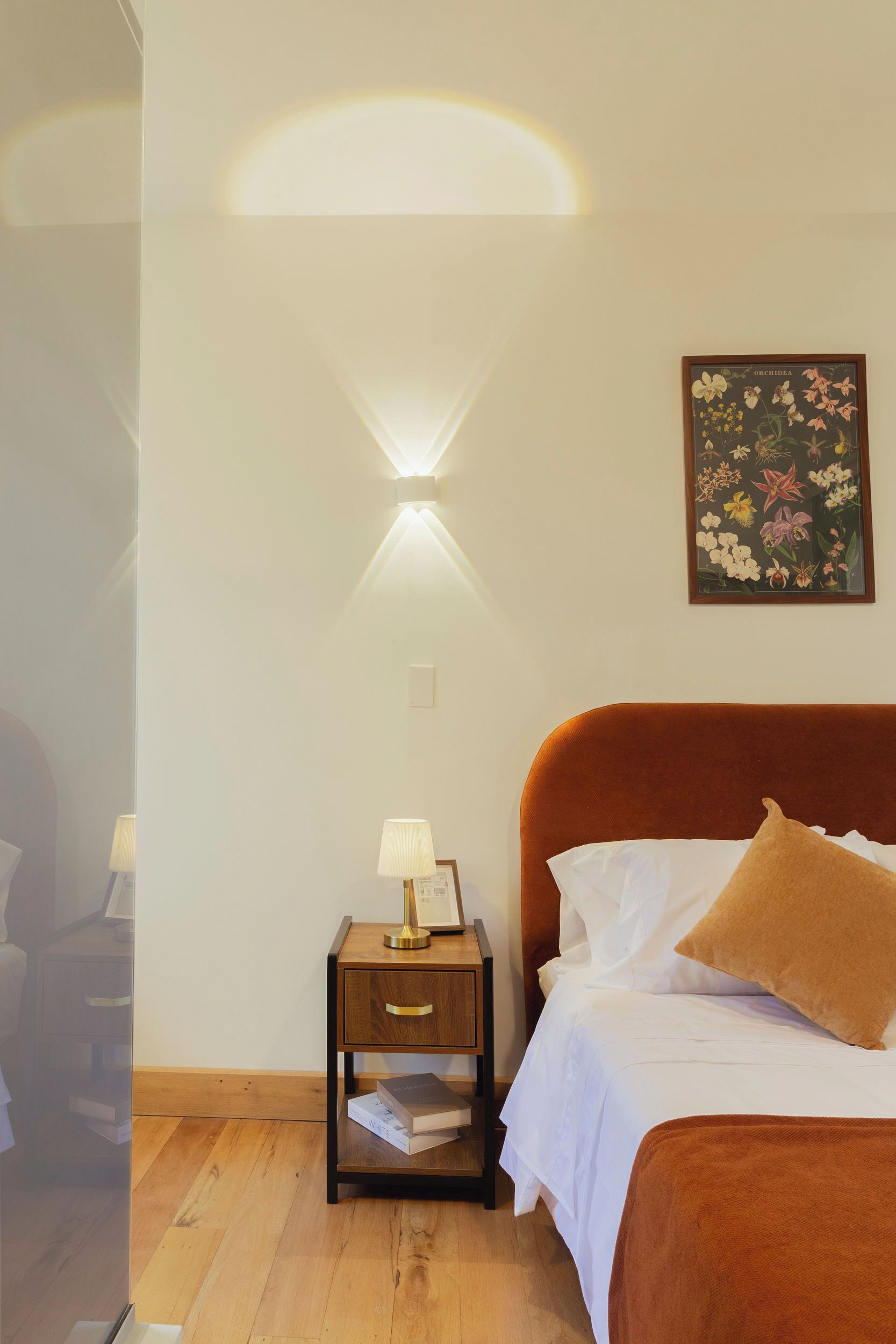 Ein Schlafzimmer mit einem Bett, einem Nachttisch mit Lampe, einer gerahmten Pflanzenillustration an der Wand und Holzboden.