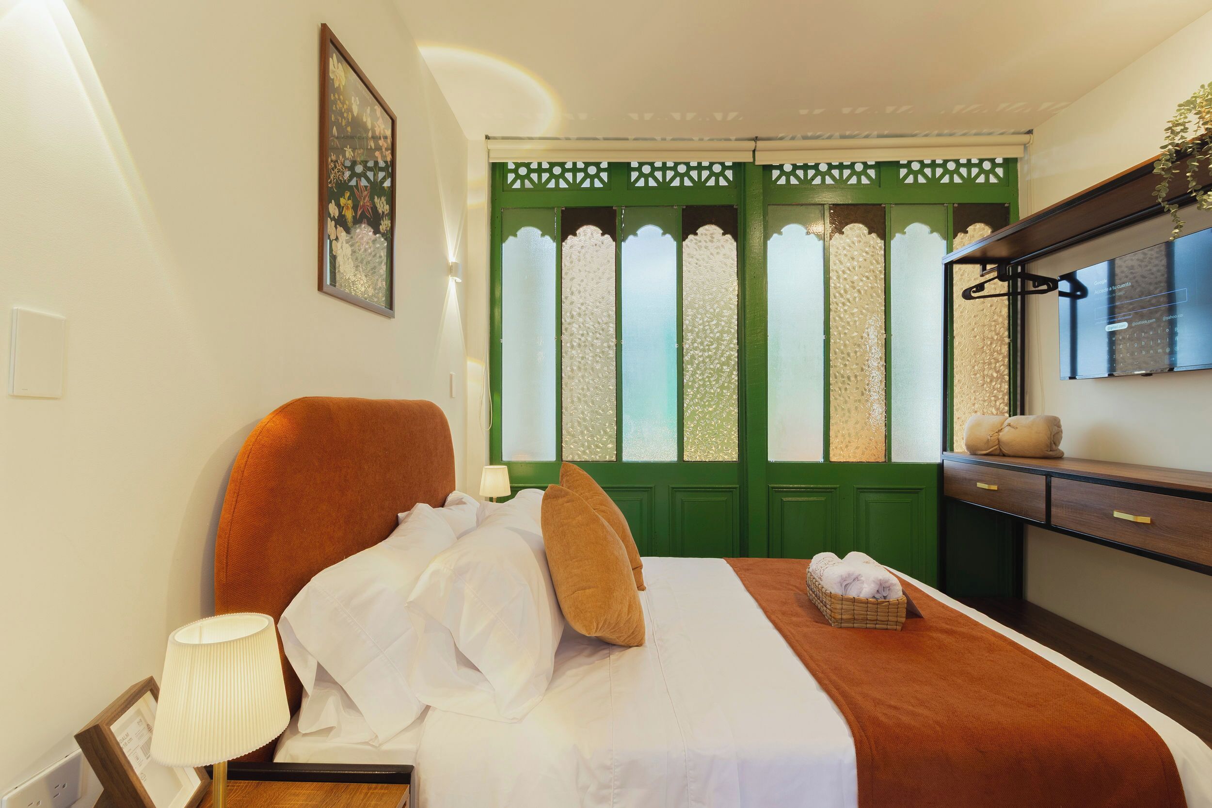 Habitación de hotel con cama, lámpara de noche, puerta verde con paneles de vidrio decorativos y cómoda de madera con espejo.