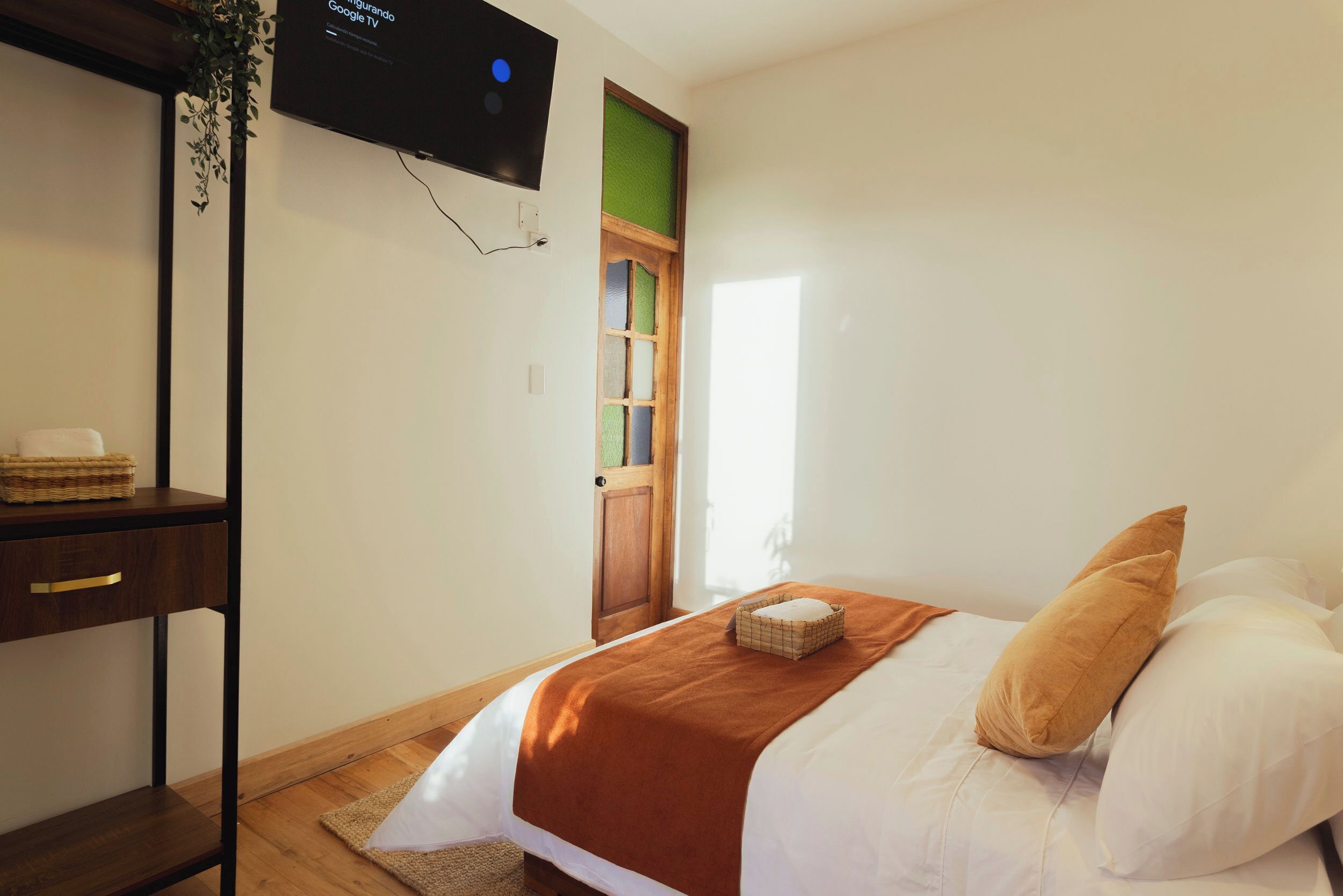 A bedroom with a bed, a TV mounted on the wall, a wooden door, and a small shelf with a basket.