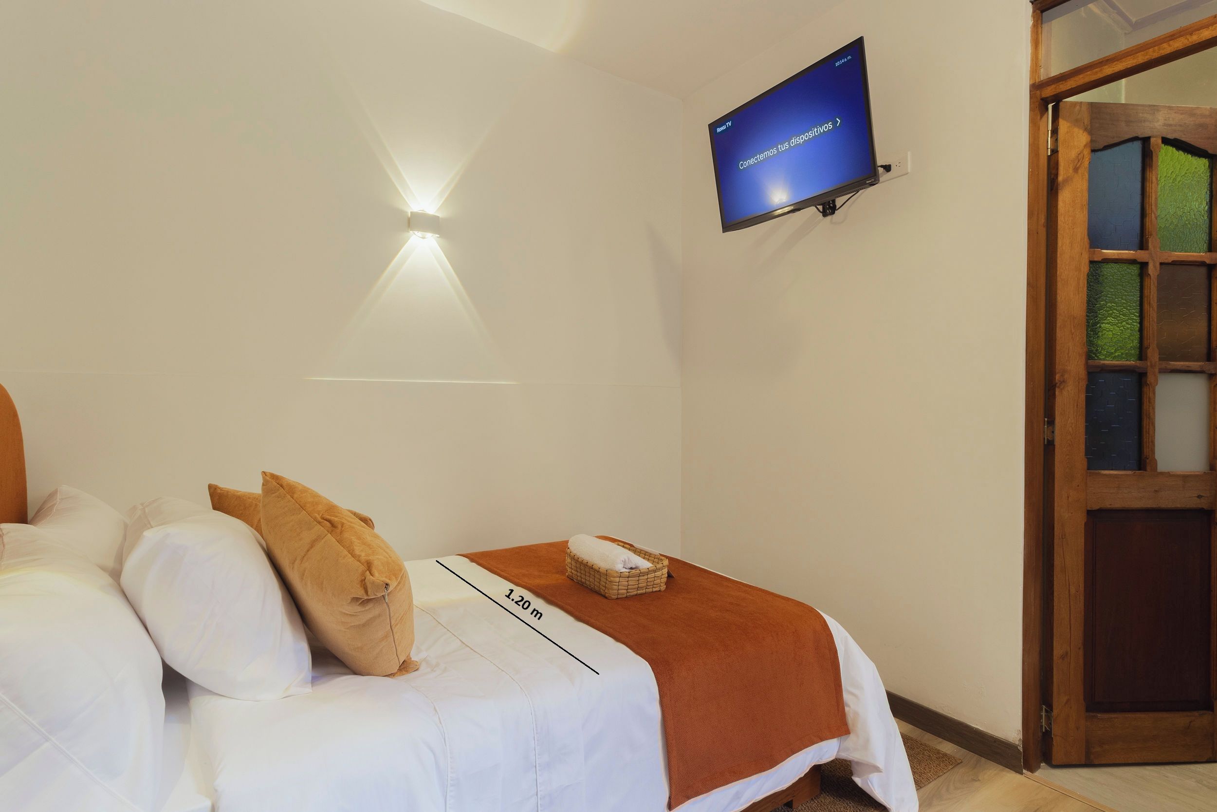 A bedroom with a bed, pillows, a bedside table with a basket, a wall-mounted TV, and a door with glass panels.
