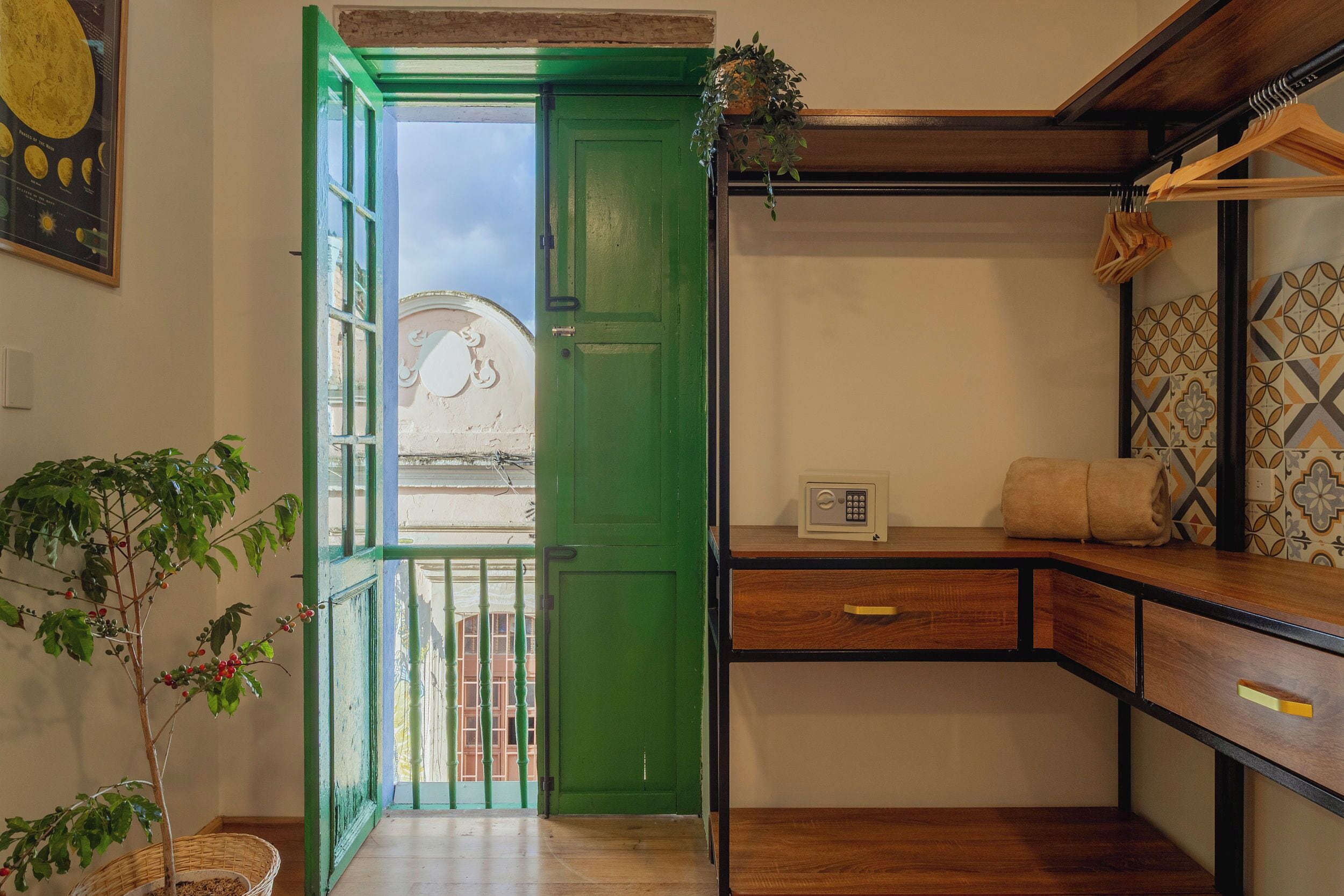 Una habitación con puerta verde, una mesita con una caja fuerte, una planta y vistas al exterior.