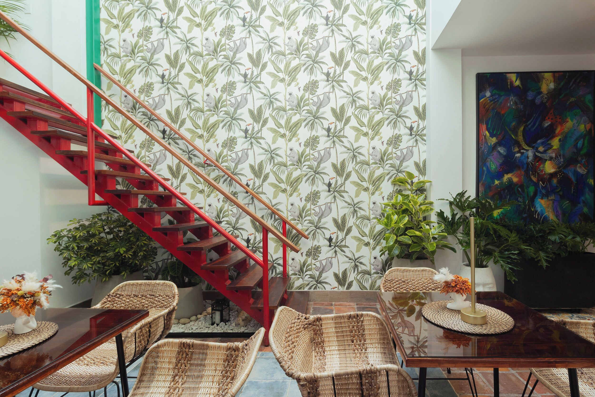 Una habitación con escalera roja, mobiliario de mimbre, un cuadro grande y un papel tapiz tropical.