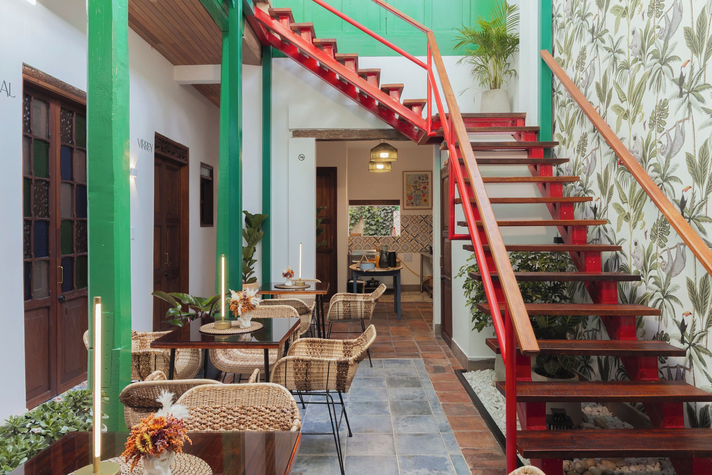 Un comedor con mobiliario de mimbre, una escalera con barandillas rojas y un papel tapiz tropical.