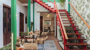 Un comedor con mobiliario de mimbre, una escalera con barandillas rojas y un papel tapiz tropical.
