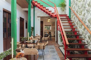Un comedor con mobiliario de mimbre, una escalera con barandillas rojas y un papel tapiz tropical.