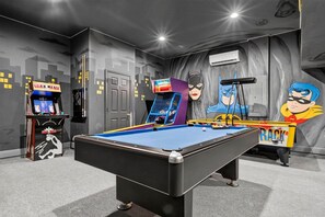 A pool table in a room with a mural of Batman and Robin, a basketball arcade machine, and a ceiling-mounted air conditioning unit. - House 7Q With Pool, Spa and Game Room (Kissimmee)