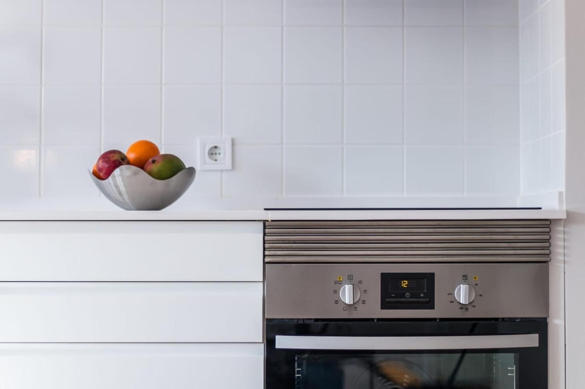 Eine Küche mit weißen Fliesen, einem Edelstahlbackofen und einer Fruchtschüssel auf der Arbeitsfläche.