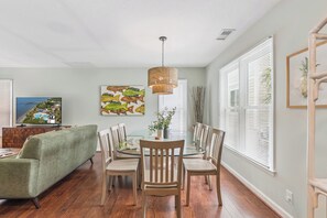 A dining area with a round glass table, wooden chairs, a woven pendant light, and a painting of fish on the wall. - Breezy Bliss located near Coast Guard Beach Entrance, sleeps 9 (St. Simons Island)