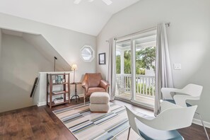 A modern living room with a brown leather chair, a white armchair, a small side table with a lamp, a bookshelf, and a large sliding glass door leading to a balcony. - Breezy Bliss located near Coast Guard Beach Entrance, sleeps 9 (St. Simons Island)