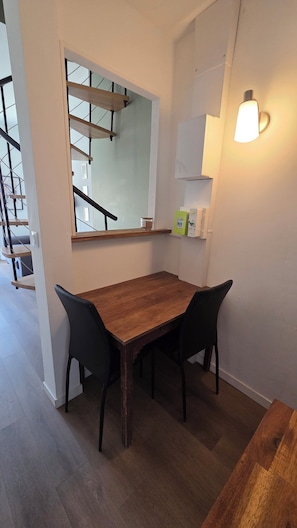 A dining area with a wooden table and black chairs, a window with a view of a staircase, and a wall-mounted light fixture. - A Cozy One-Bedroom Apartment in a Charming, Authentic Village (Saint-Fort-sur-Gironde)