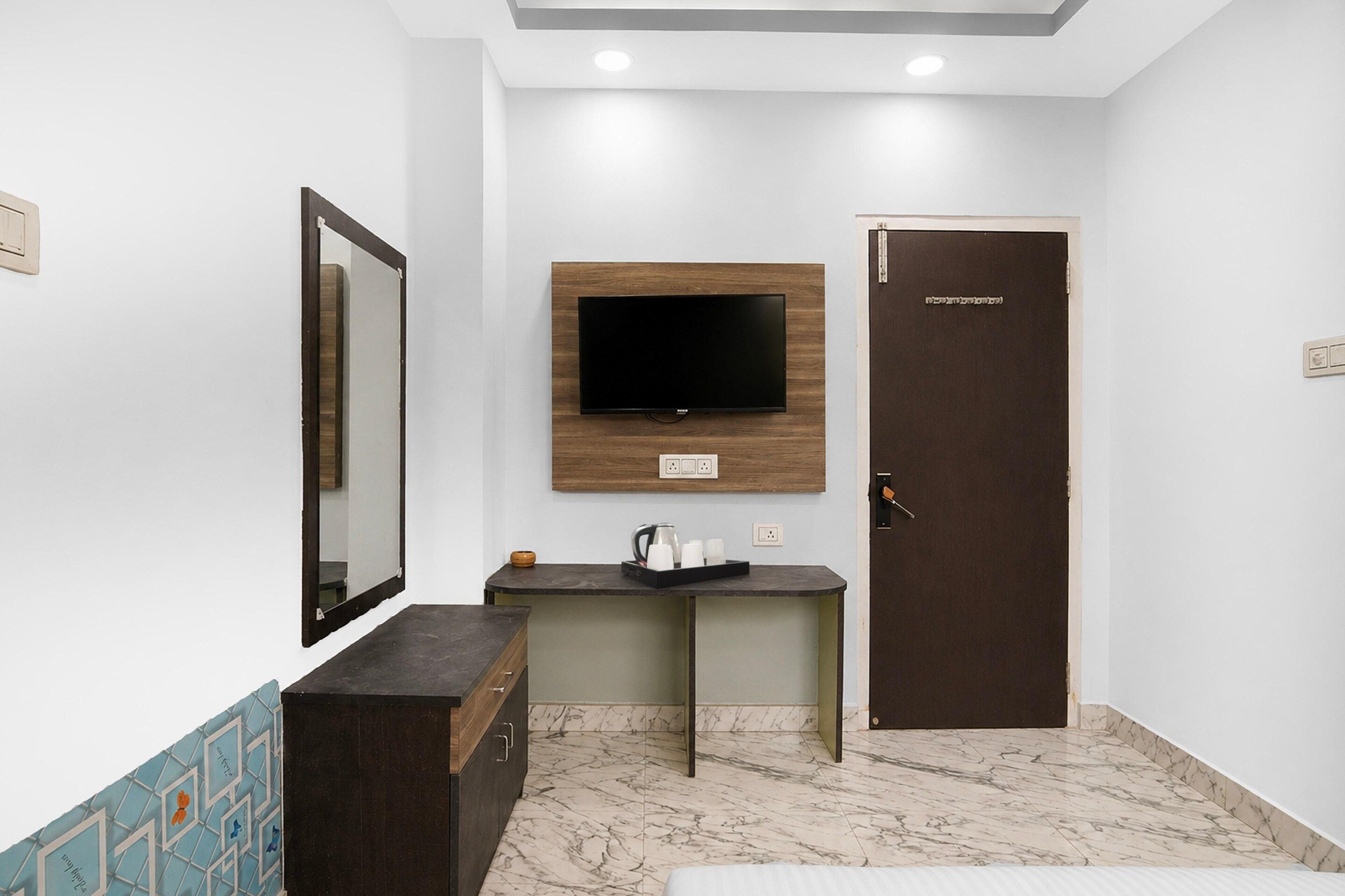 Ein Hotelzimmer mit Flachbildfernseher an der Wand, einer Holztür, einem kleinen Schreibtisch mit Wasserkocher und einem Spiegel.